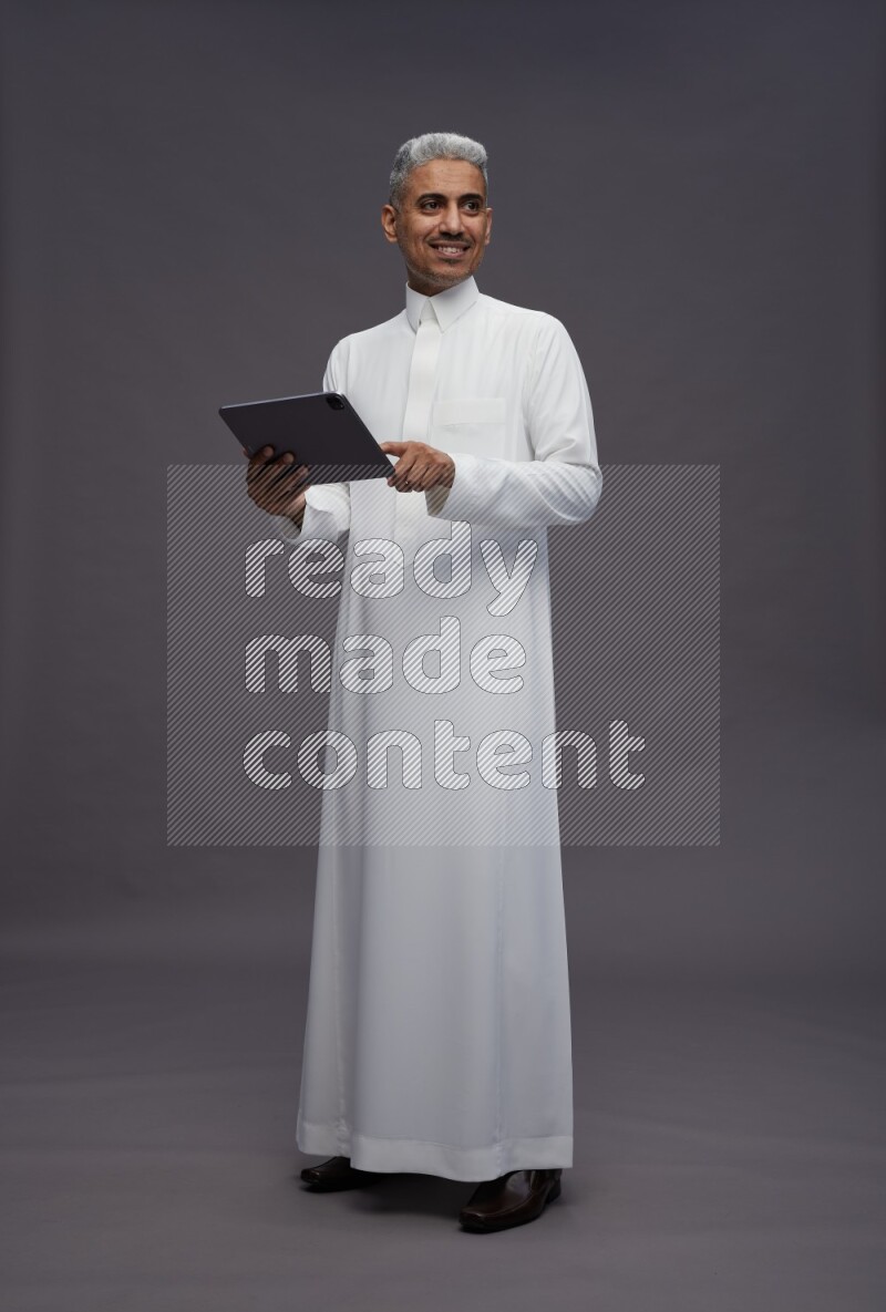 Saudi man wearing thob standing working on tablet on gray background