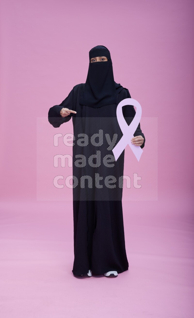 Saudi woman wearing abaya and niqab standing holding awareness ribbon on pink background