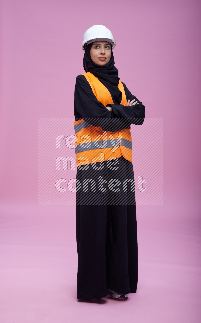 Saudi woman wearing Abaya with engineer vest and helmet standing with crossed arms on pink background