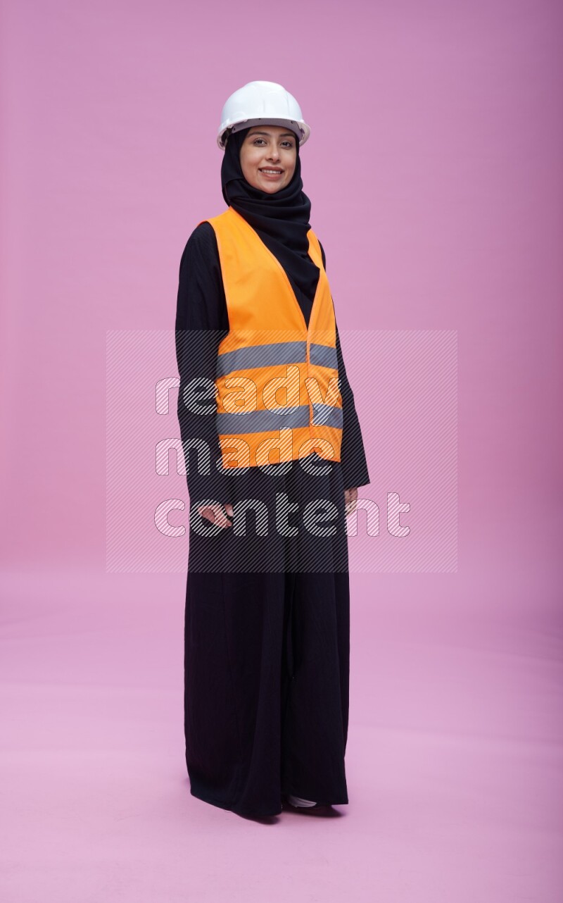 Saudi woman wearing Abaya with engineer vest and helmet standing interacting with the camera on pink background