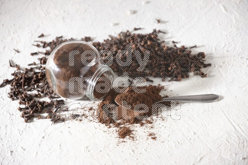 A flipped glass spice jar and a metal spoon full of cloves powder and powder came out of the jar with cloves spread on textured white flooring