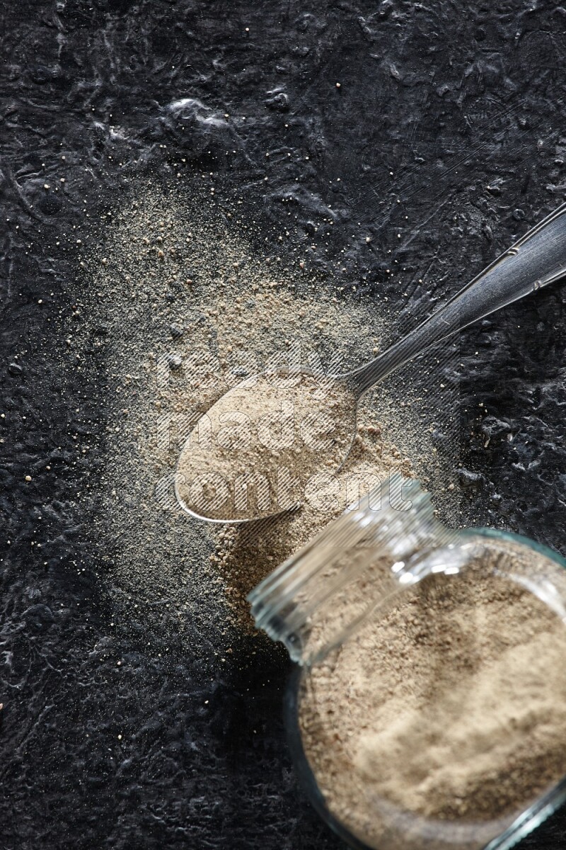 A flipped herbal glass jar and a metal spoon full of white pepper powder with spilled powder on textured black flooring