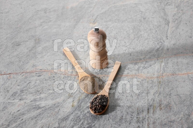 ملعقتان خشبيتان، ملعقة ممتلئة بمسحوق الفلفل الأسود والأخرى ممتلئة بحبات الفلفل الأسود ومطحنة خشبية على خلفية رخام رمادية