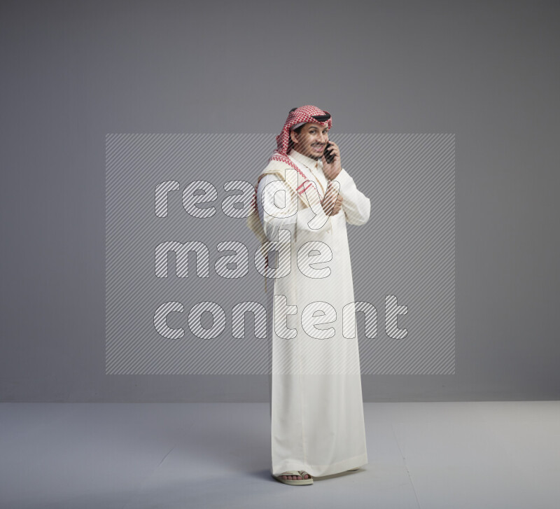 A Saudi man standing wearing thob and red shomag talking on phone on gray background