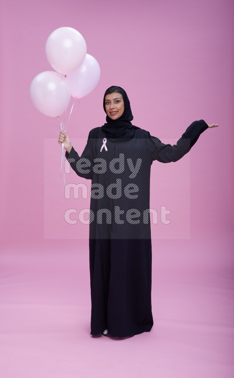 Saudi woman wearing pink ribbon on Abaya standing holding balloons on plain pink background