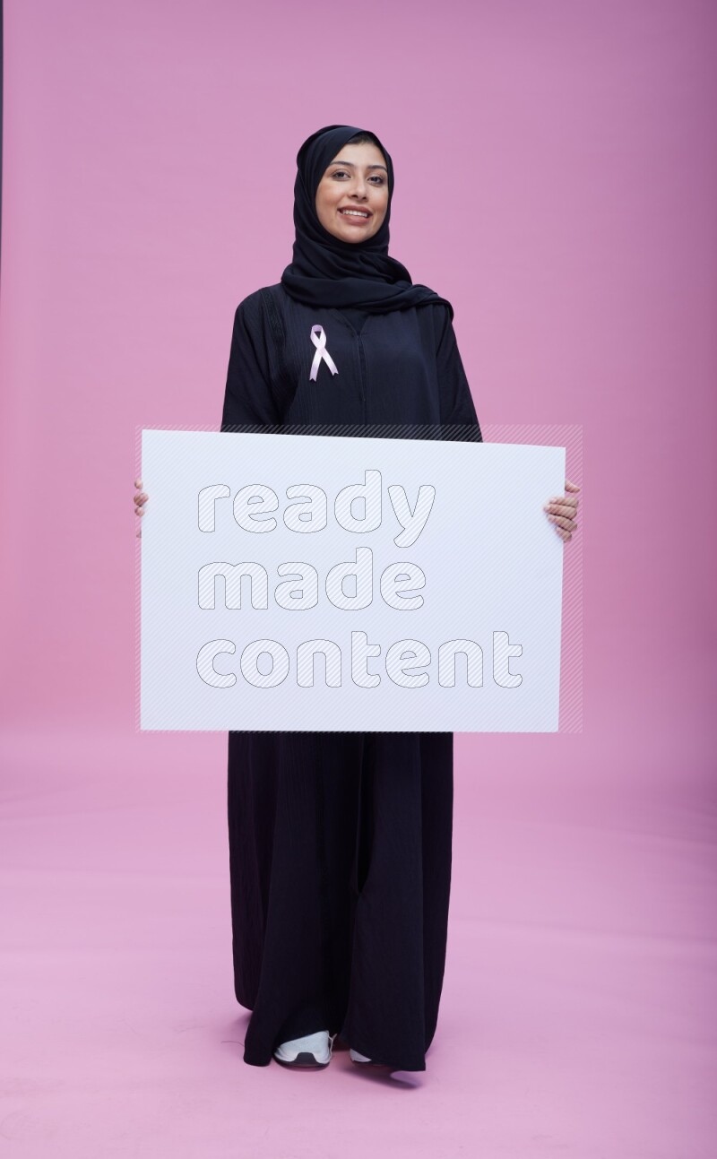 Saudi woman wearing pink ribbon on Abaya standing holding board on pink background