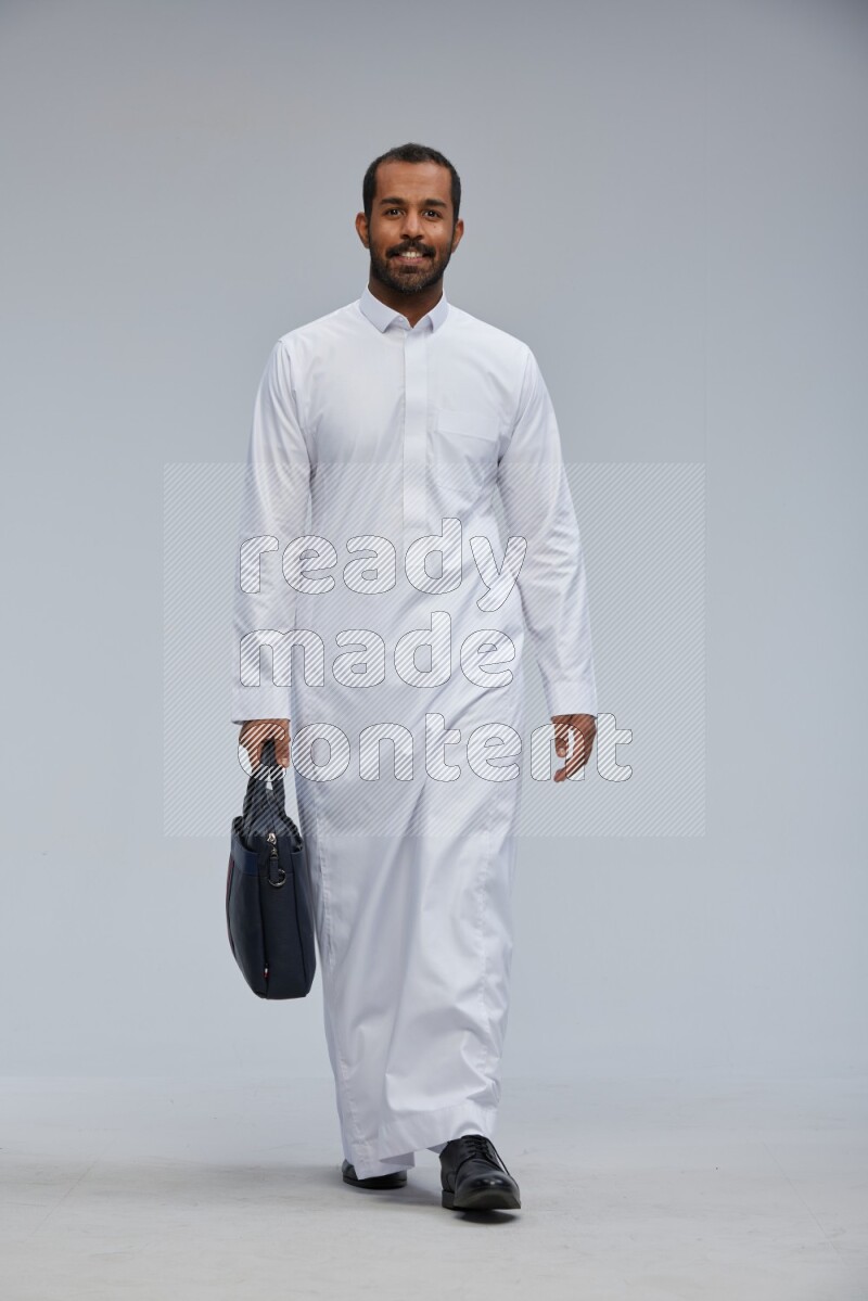 Saudi man wearing thob standing holding suitcase on gray background