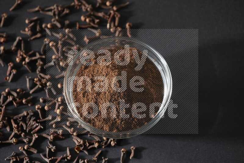 وعاء زجاجي ممتلئ ببودرة القرنفل مع حبوب القرنفل المتناثرة على أرضية سوداء