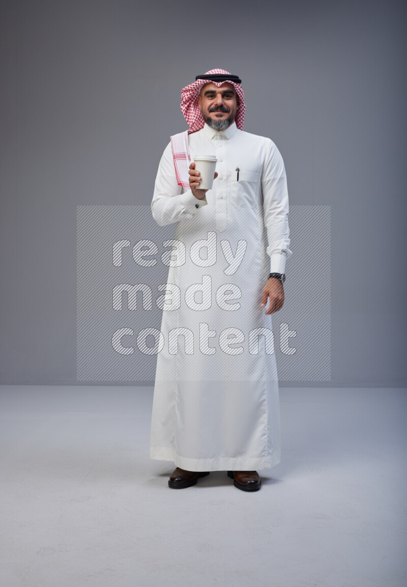 Saudi man Wearing Thob and red Shomag standing holding paper cup on Gray background