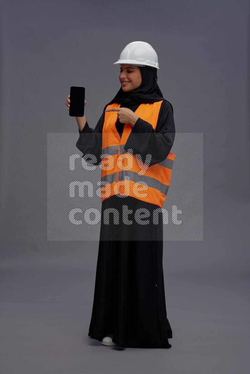Saudi woman wearing Abaya with engineer vest standing showing phone to camera on gray background