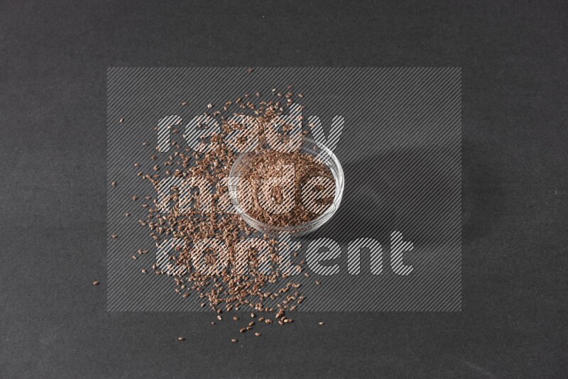 A glass bowl full of flaxseeds surrounded by the seeds on a black flooring
