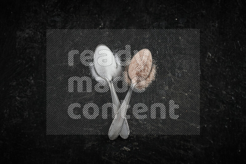2 metal spoons filled with white salt and pink himalayan salt on black background
