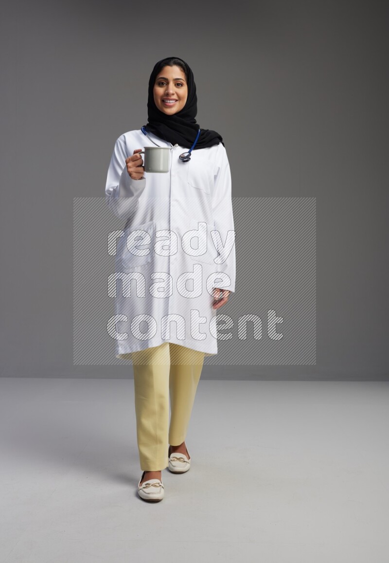 Saudi woman wearing lab coat with stethoscope standing holding mug on Gray background