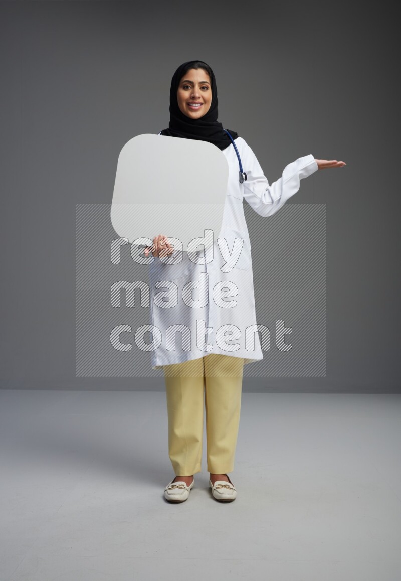 Saudi woman wearing lab coat with stethoscope standing holding social media sign on Gray background