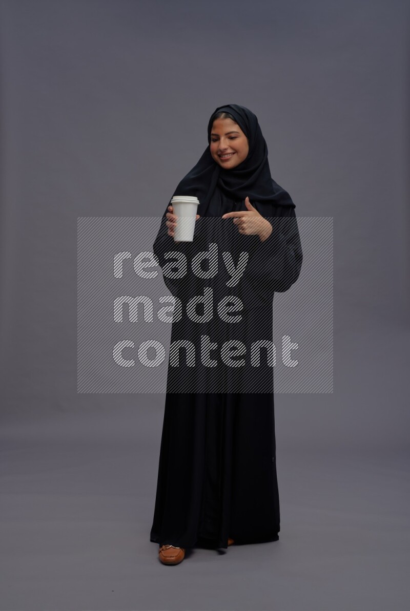 Saudi woman wearing Abaya standing holding paper cup on gray background