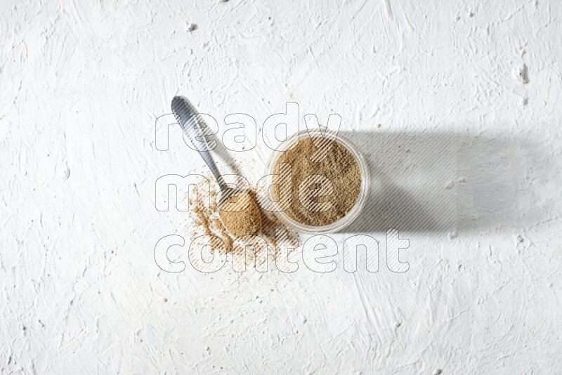 A glass jar and a metal spoon full of cumin powder on textured white flooring