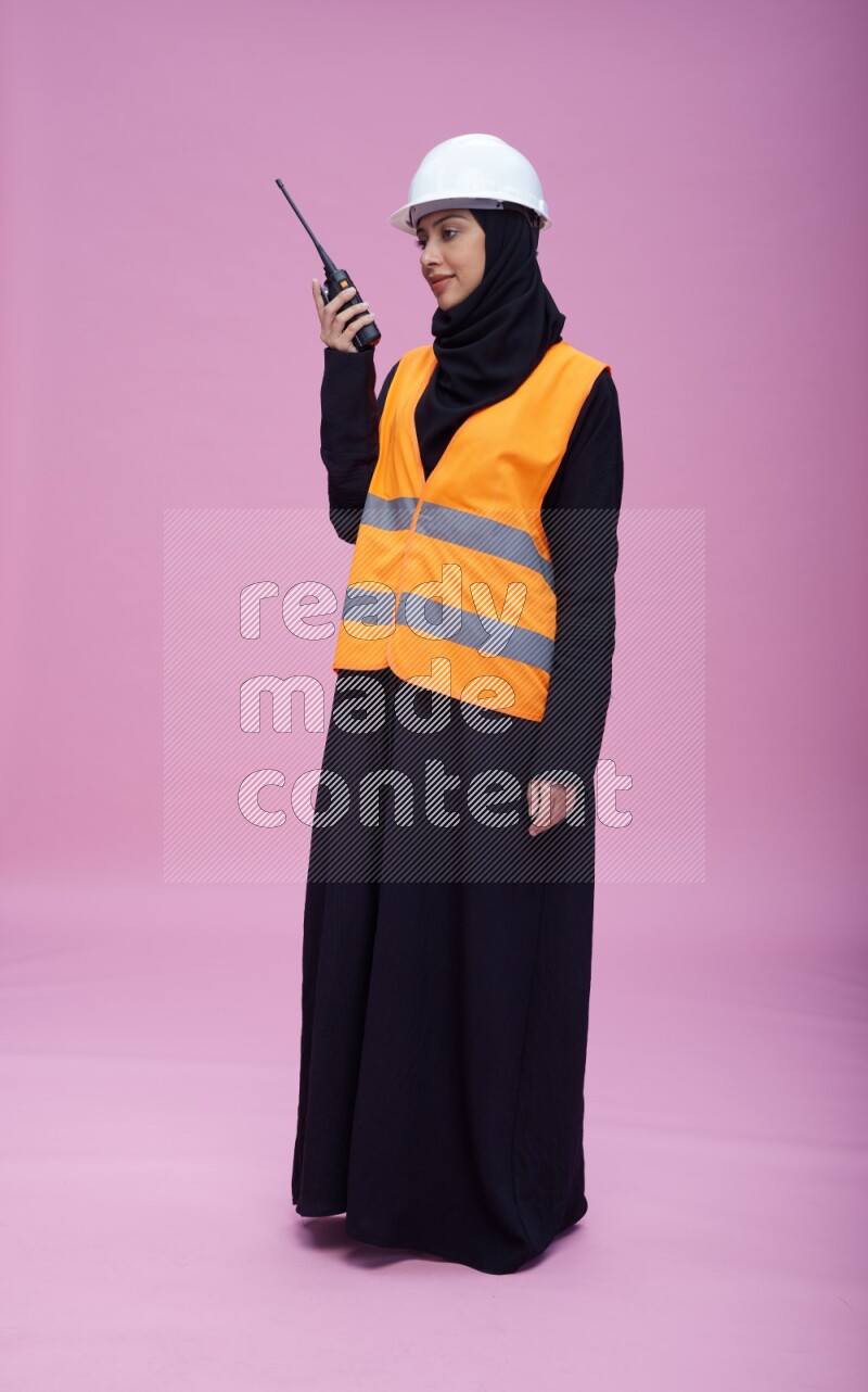 Saudi woman wearing Abaya with engineer vest and helmet standing holding walkie-talkie on pink background