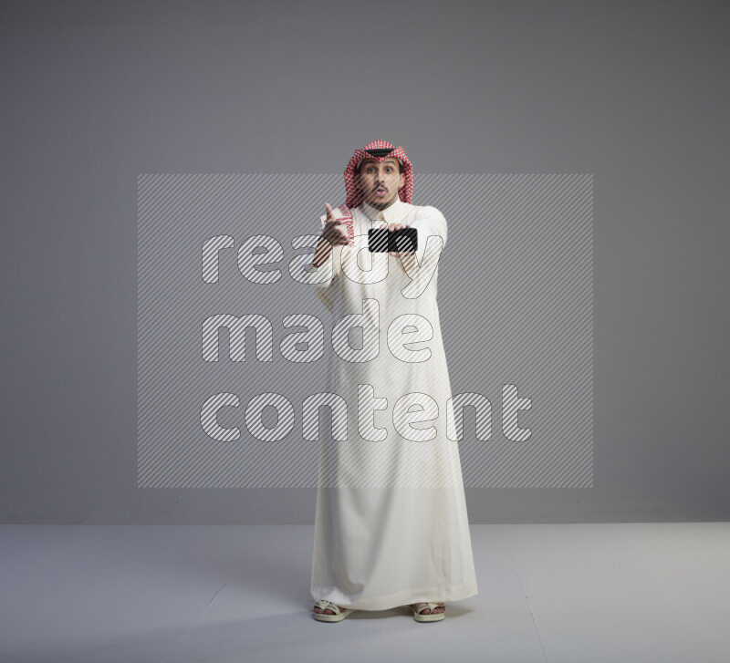 A Saudi man standing wearing thob and red shomag showing phone to camera on gray background