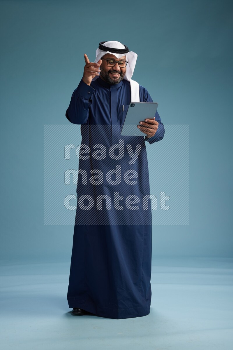 Saudi Man with shimag Standing holding ATM card while working on tablet on blue background