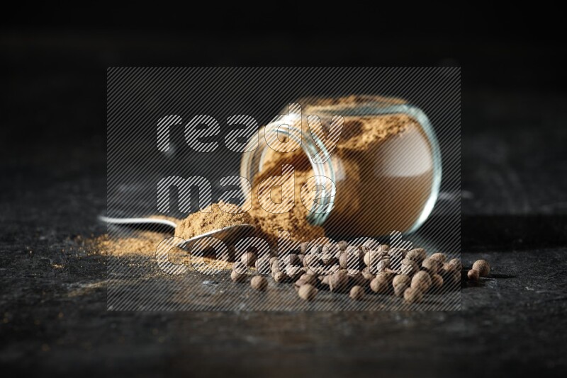 وعاء زجاجي للتوابل مقلوب وملعقة معدنية ممتلئان ببودرة بهار الفلفل الحلو والبودرة تناثرت من الوعاء مع بعض الحبوب على خلفية سوداء