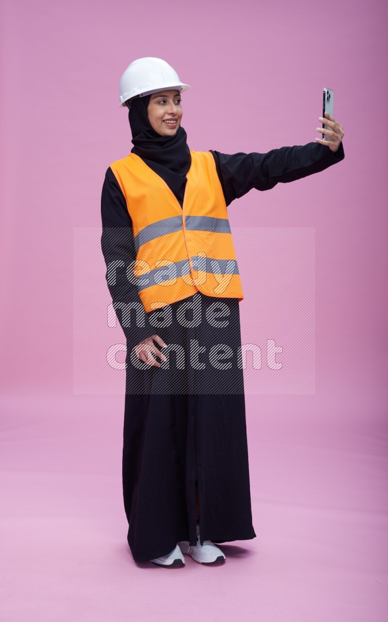 Saudi woman wearing Abaya with engineer vest and helmet standing taking selfie on pink background