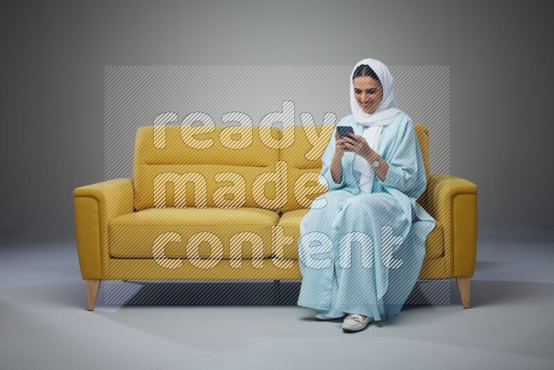 A Saudi woman wearing a light blue Abaya and white head scarf sitting on a yellow sofa and using her phone eye level on a grey background