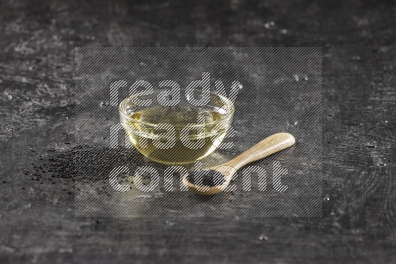 A glass bowl full of black seeds oil and wooden spoon full of black seeds with seeds spreaded on a textured black flooring