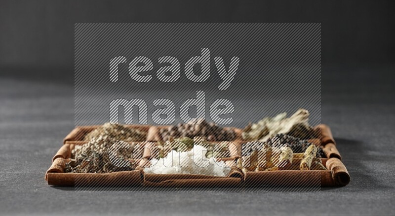 9 squares of cinnamon sticks full of cardamom in the middle surrounded by turmeric fingers, black pepper, bay laurel leaves, allspice, cumin, white pepper, dried basil and salt on black flooring