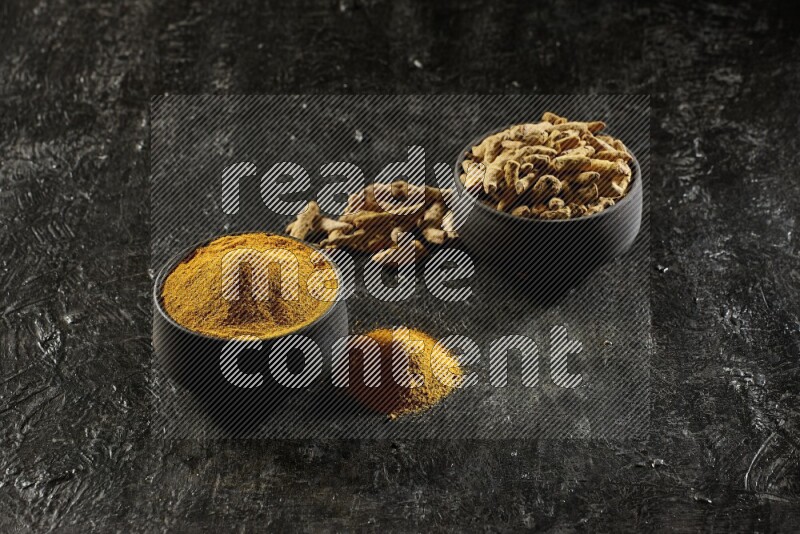 2 pottery black bowls, one full of turmeric powder and the other full of dried turmeric whole fingers on a textured black flooring