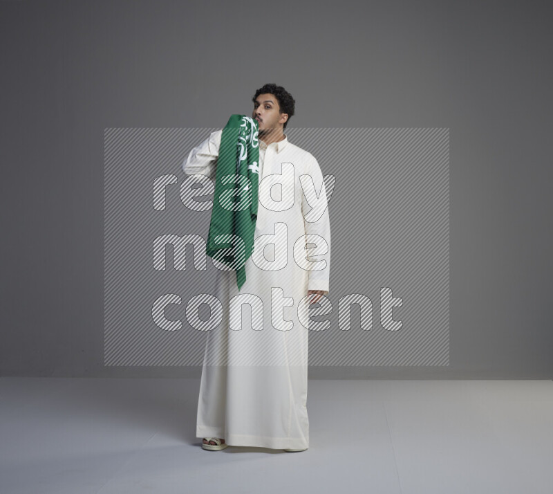 A Saudi man standing wearing thob kissing Saudi flag on gray background