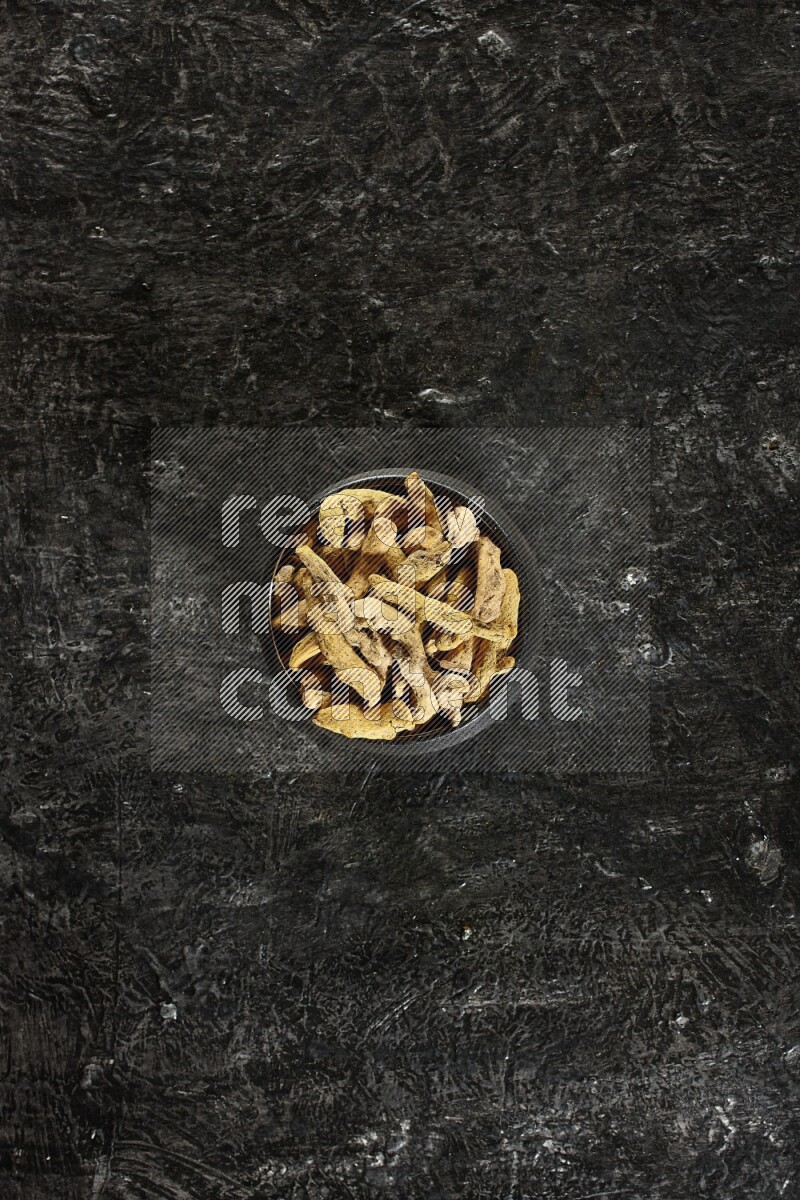 A black pottery bowl full of dried turmeric whole fingers on textured black flooring