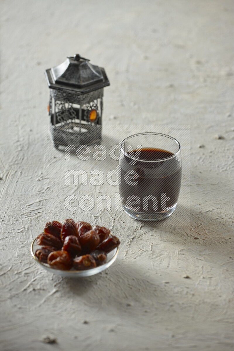 A silver lantern with different drinks, dates, nuts, prayer beads and quran on textured white background