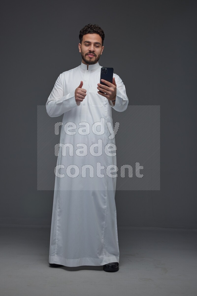 A Saudi man wearing Thobe and making a video call using his phone while standing and making different poses eye level on a gray background