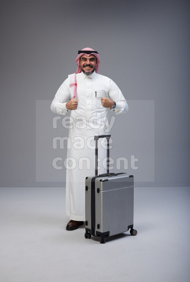 Saudi man wearing Thob and red Shomag standing holding Travel bag on Gray background