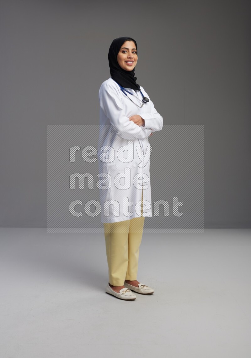 Saudi woman wearing lab coat with stethoscope standing with crossed arms on Gray background