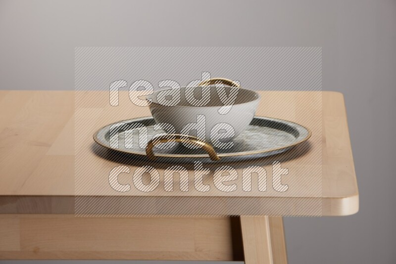 off white bowl with islamic pattern placed on a rounded stainless steel tray with golden handels on the edge of wooden table
