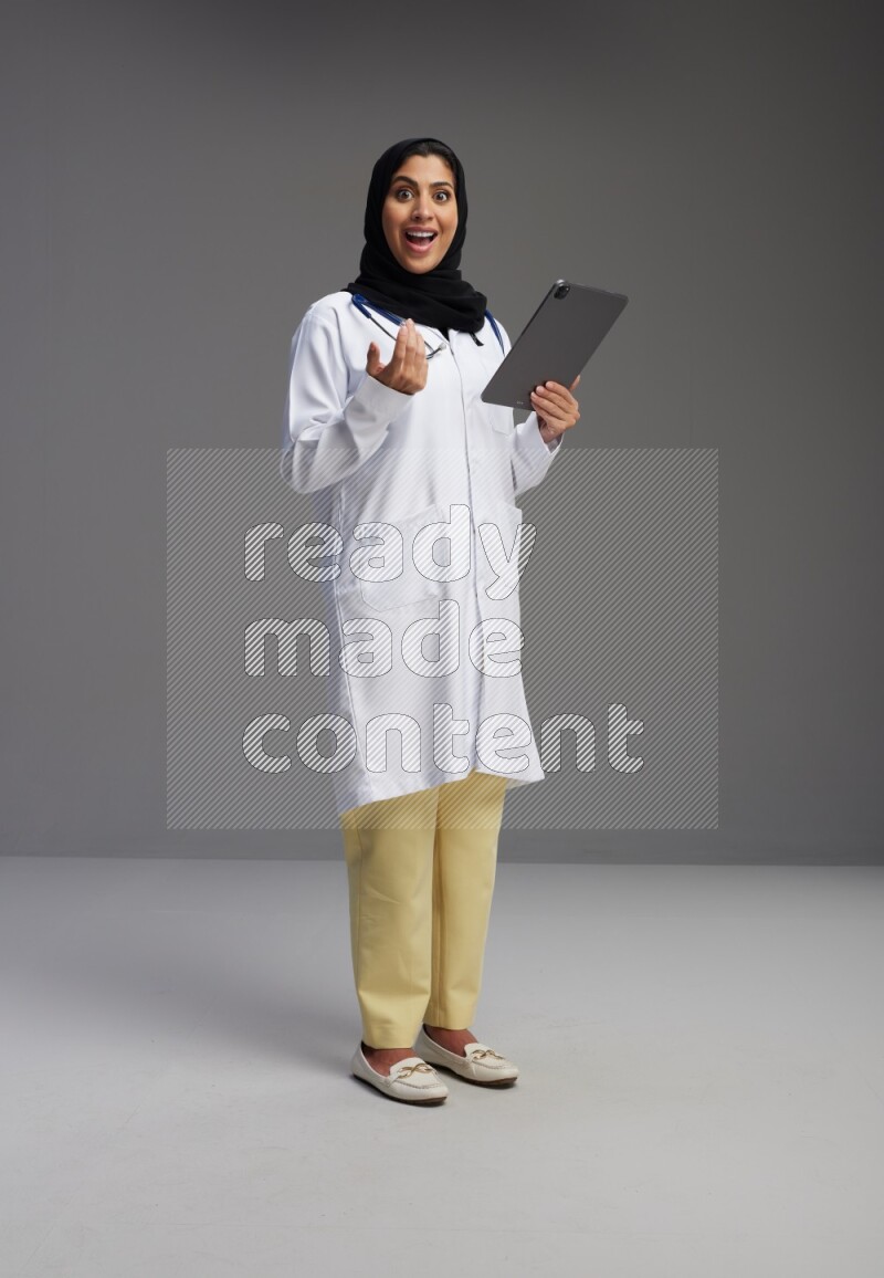 Saudi woman wearing lab coat with stethoscope standing working on tablet on Gray background