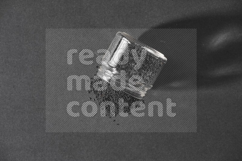 وعاء زجاجي مقلوب ممتلئ ببذور حبة البركة والبذور متناثرة على أرضية سوداء