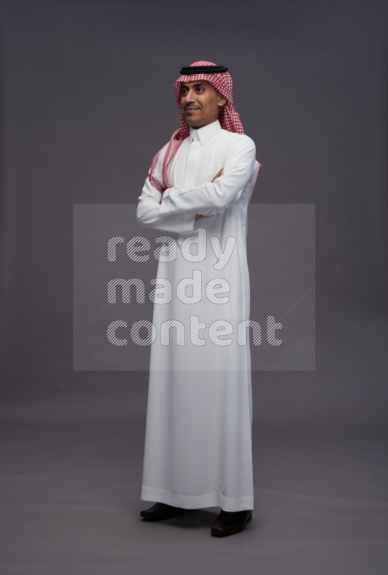 Saudi man wearing thob and shomag standing with crossed arms on gray background