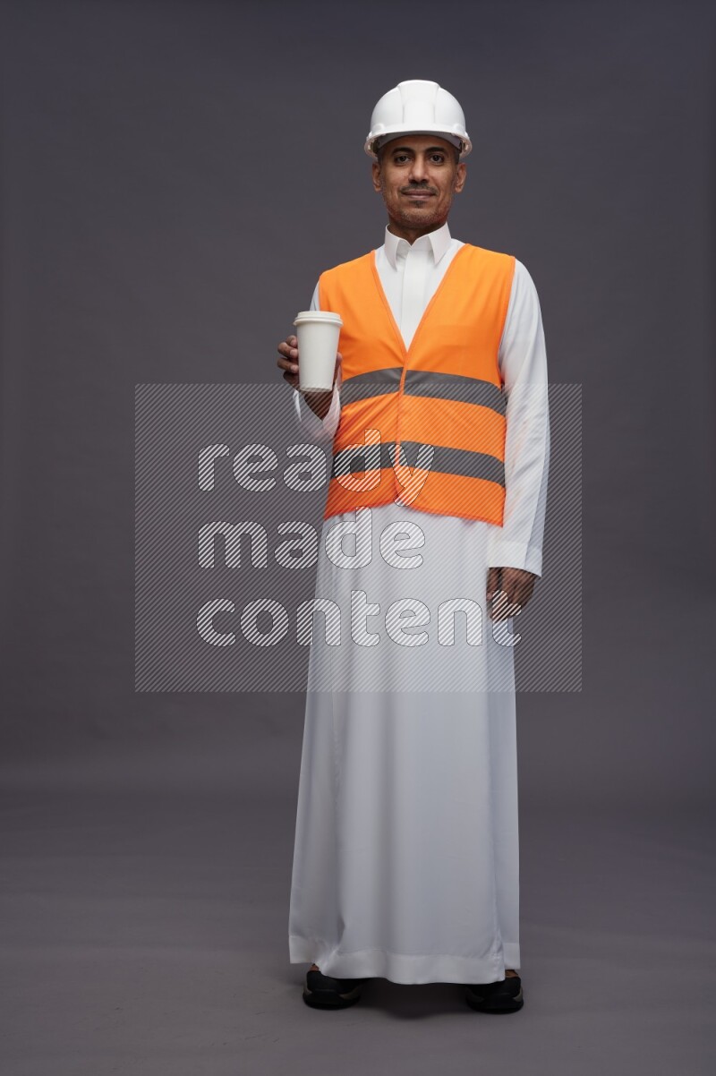 Saudi man wearing thob with engineer vest standing holding paper cup on gray background