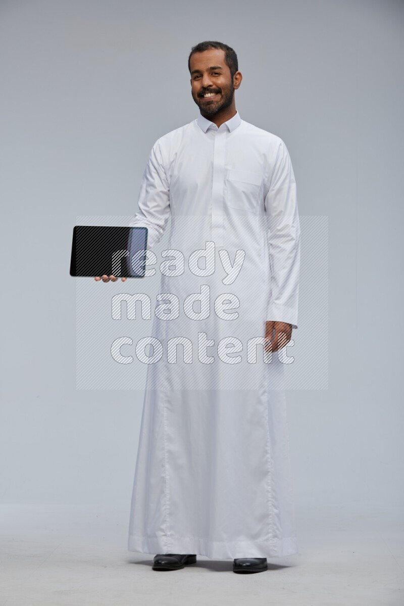 Saudi man wearing Thob standing showing tablet to camera on Gray background