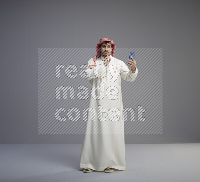 A Saudi man standing wearing thob and red shomag taking selfie on gray background