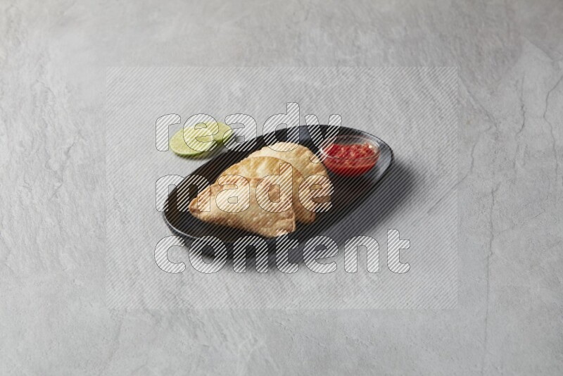 Three fried sambosas in an oval shaped black plate and a red sauce in a black round ramekin on a gray background