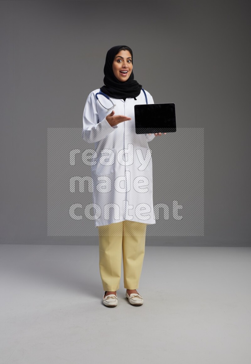 Saudi woman wearing lab coat with stethoscope standing showing tablet to camera with sign in the back on Gray background