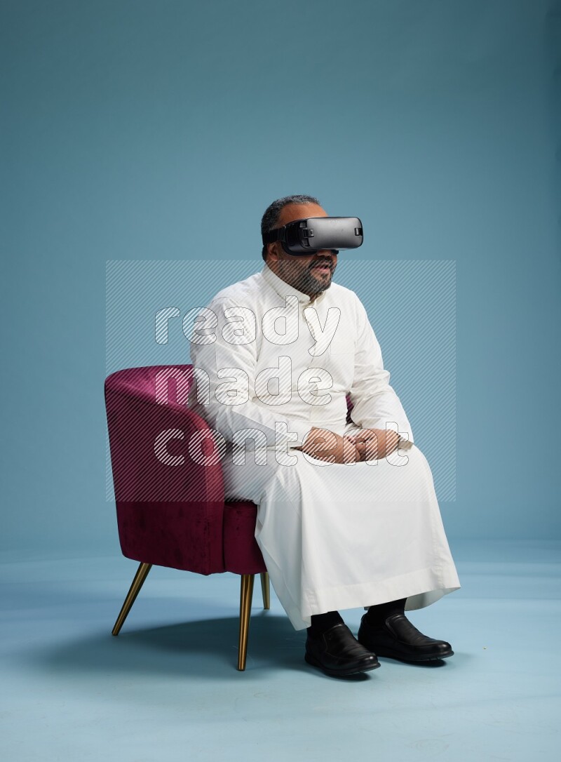 Saudi Man without shimag sitting on chair  gaming with VR on blue background