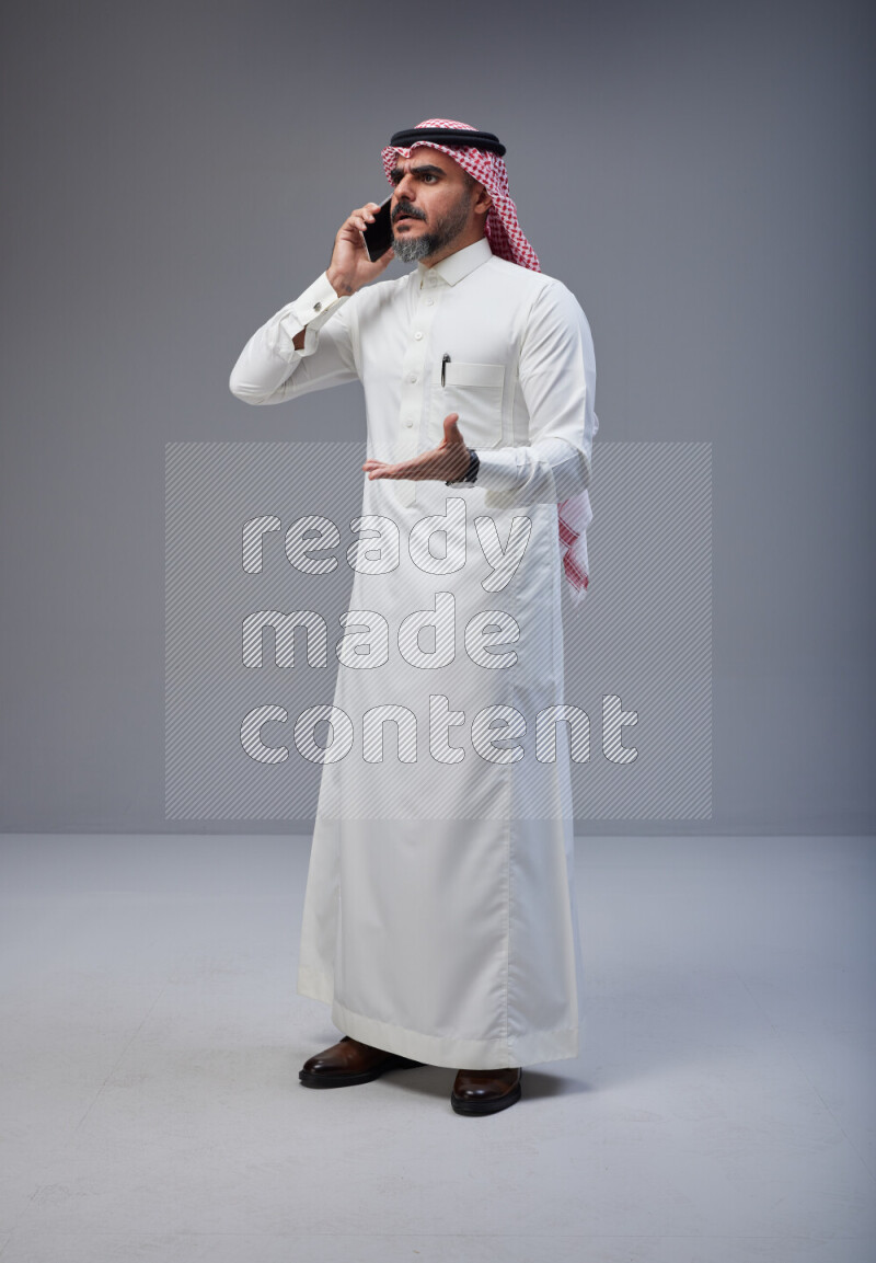 Saudi man Wearing Thob and red Shomag standing talking on phone on Gray background