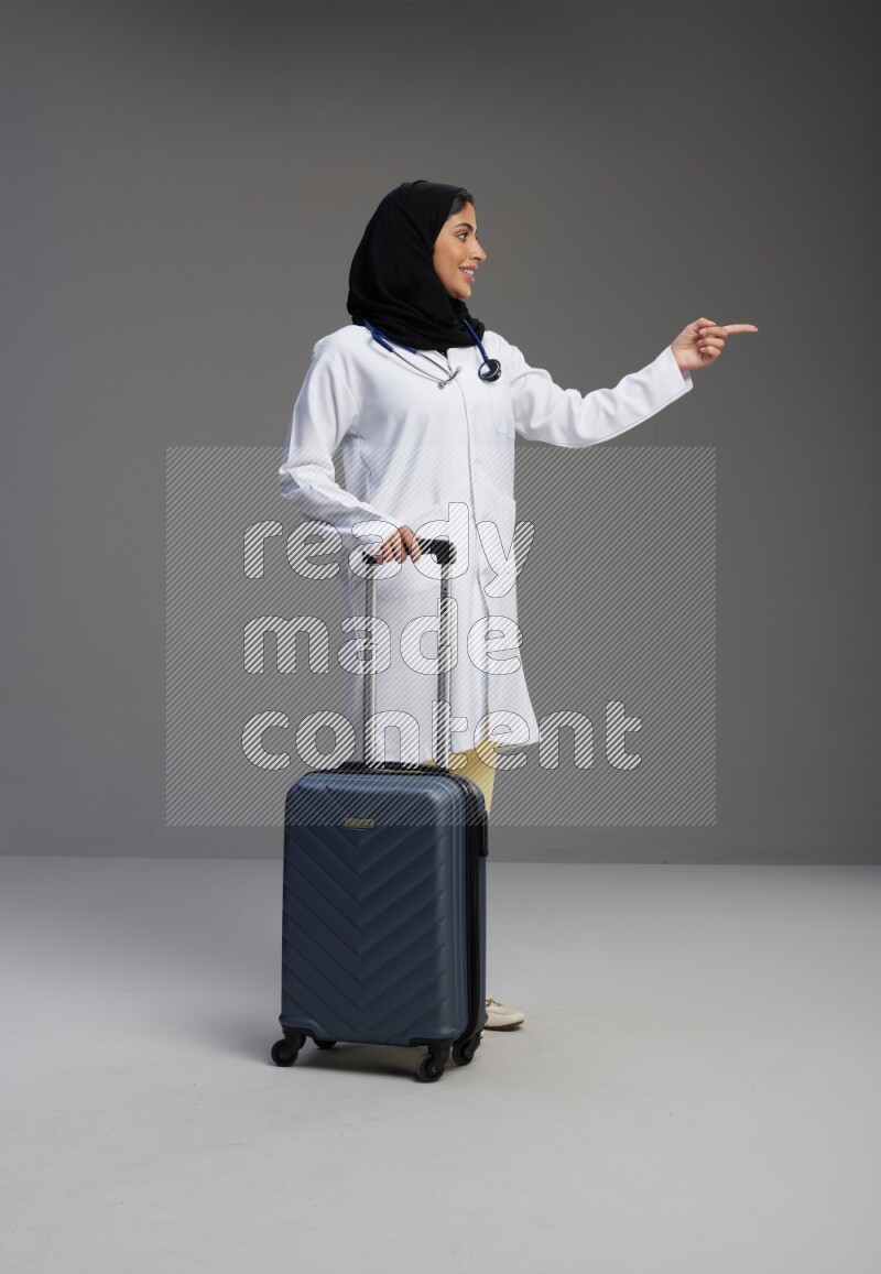 Saudi woman wearing lab coat with stethoscope standing holding Travel bag on Gray background