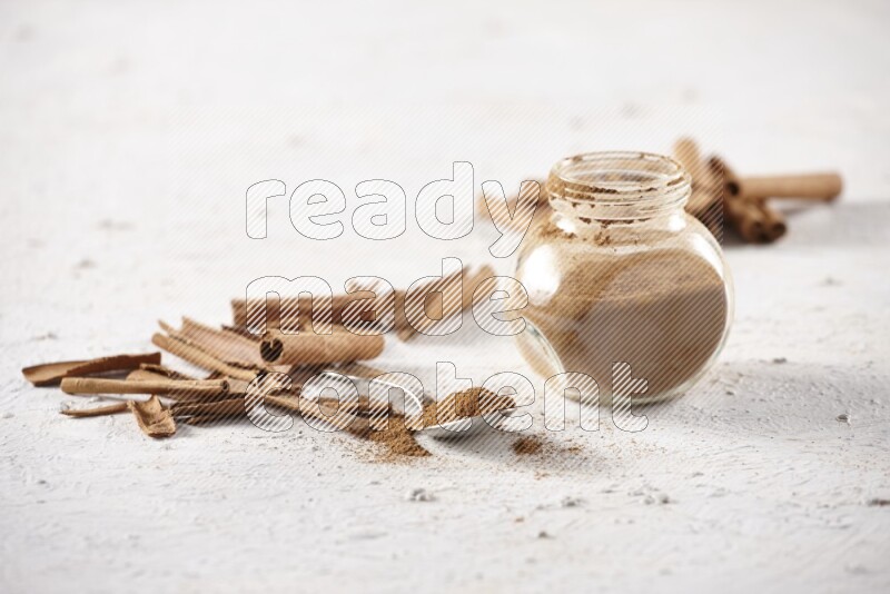 Herbal glass jar full cinnamon powder and a metal spoon surrounded by cinnamon sticks on a white background