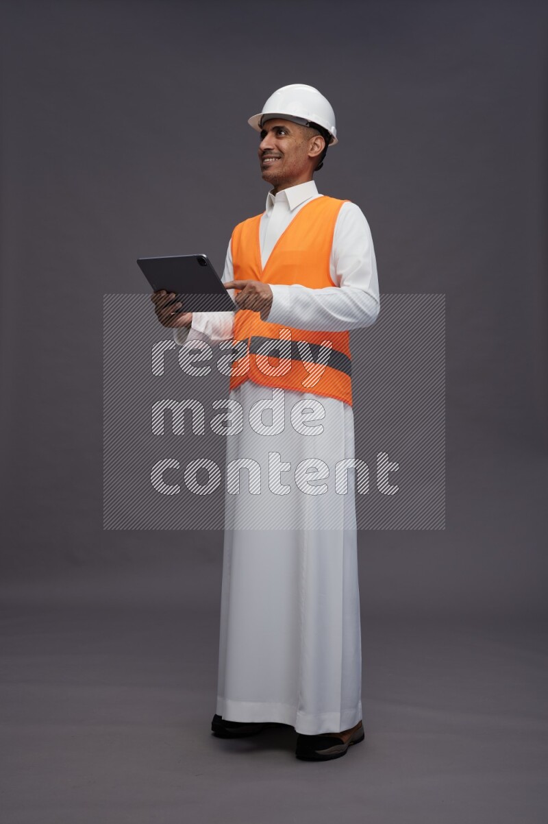 Saudi man wearing thob with engineer vest standing working on tablet on gray background