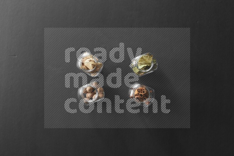 4 glass spice jars full of laurel bay leaves, ginger, cinnamon and nutmeg on black flooring